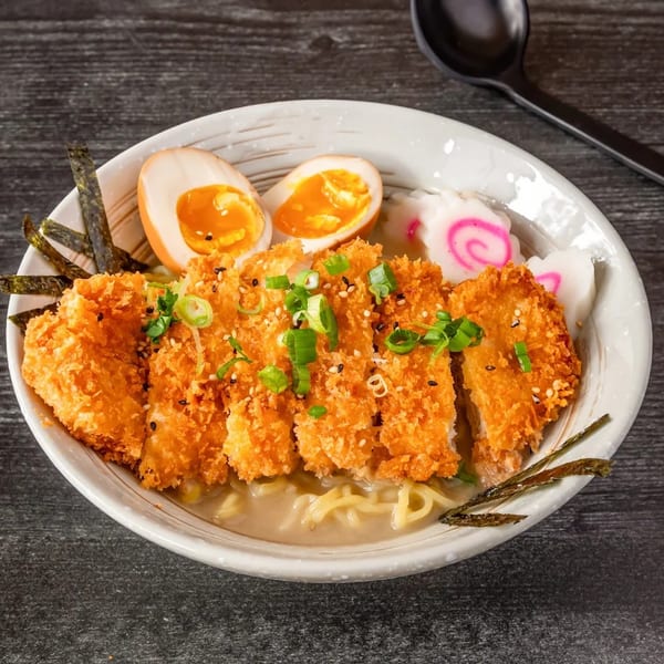 Crispy chicken katsu ramen with eggs, fish cakes, and seaweed in a white bowl.