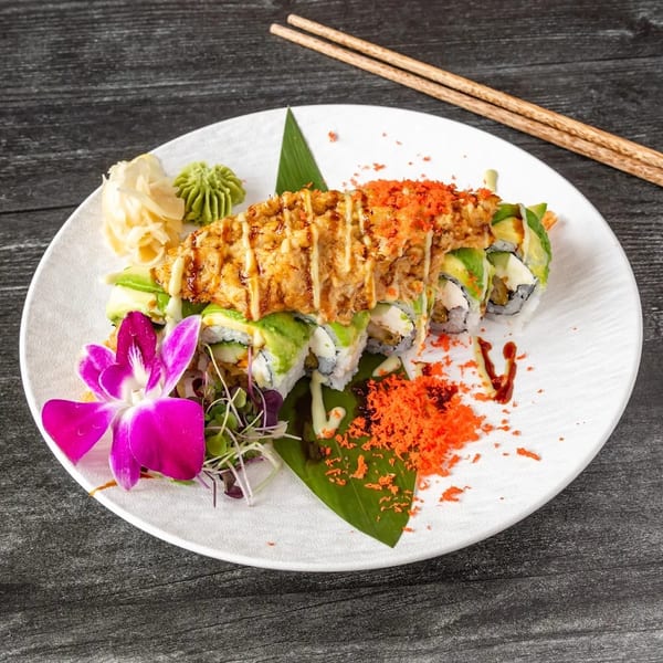Sushi roll topped with fried tempura, avocado, and orange roe, garnished with a purple orchid.