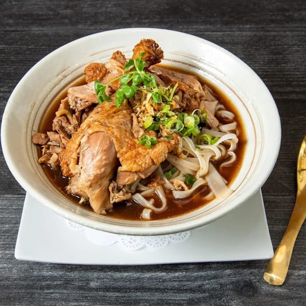 A bowl of duck noodle soup with flat rice noodles, green onions, and crispy garlic.