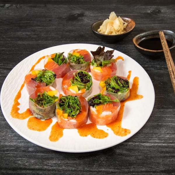 Colorful fresh spring rolls with salmon, tuna, mango, and greens on a white plate with sauce.
