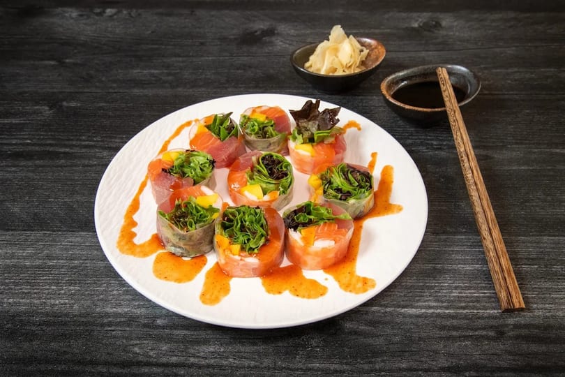 Colorful fresh spring rolls with salmon, tuna, mango, and greens on a white plate with sauce.