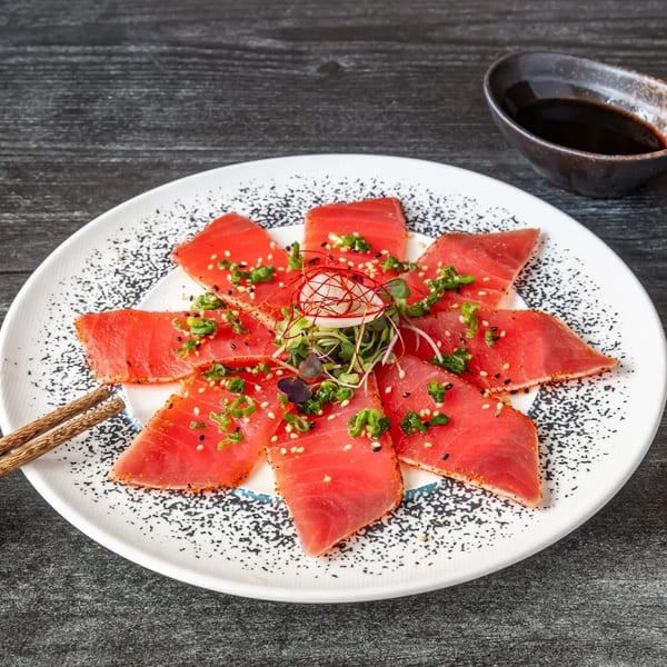 Tuna tataki with green onions, sesame seeds, and microgreens, served with chopsticks and soy sauce.