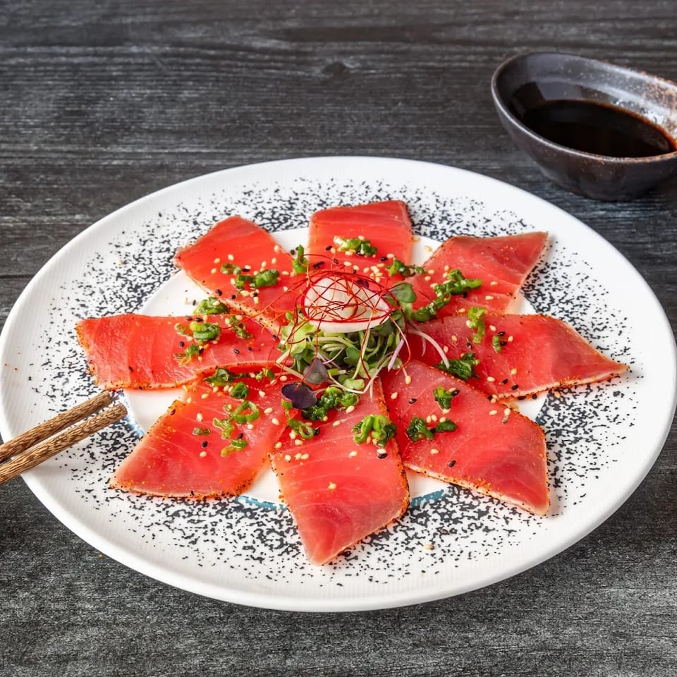 Tuna tataki with green onions, sesame seeds, and microgreens, served with chopsticks and soy sauce.