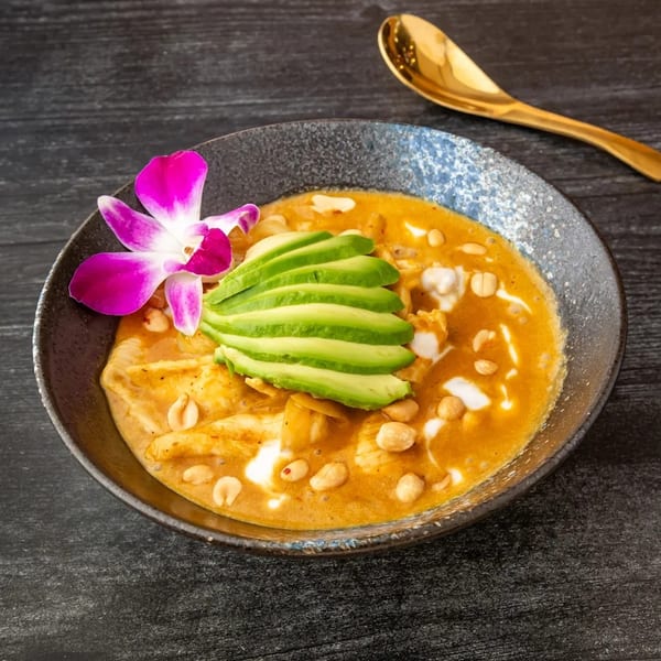 A bowl of orange curry with chicken, peanuts, avocado slices, and a purple orchid, with a gold spoon.