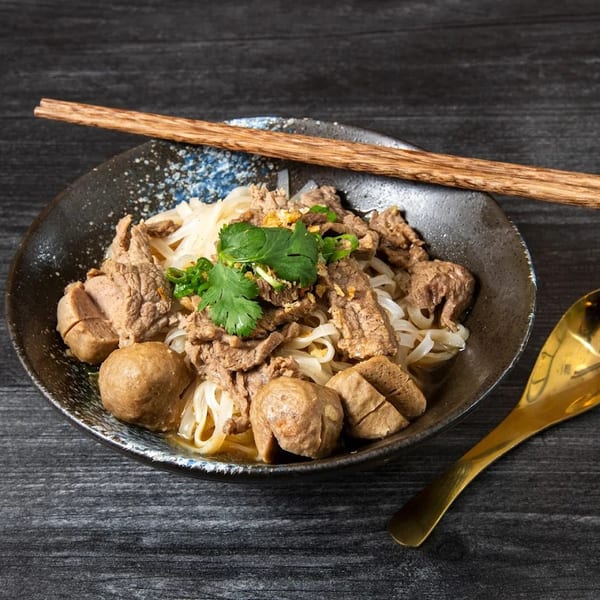A bowl of Thai boat noodles with beef, meatballs, and cilantro, with chopsticks and a spoon.