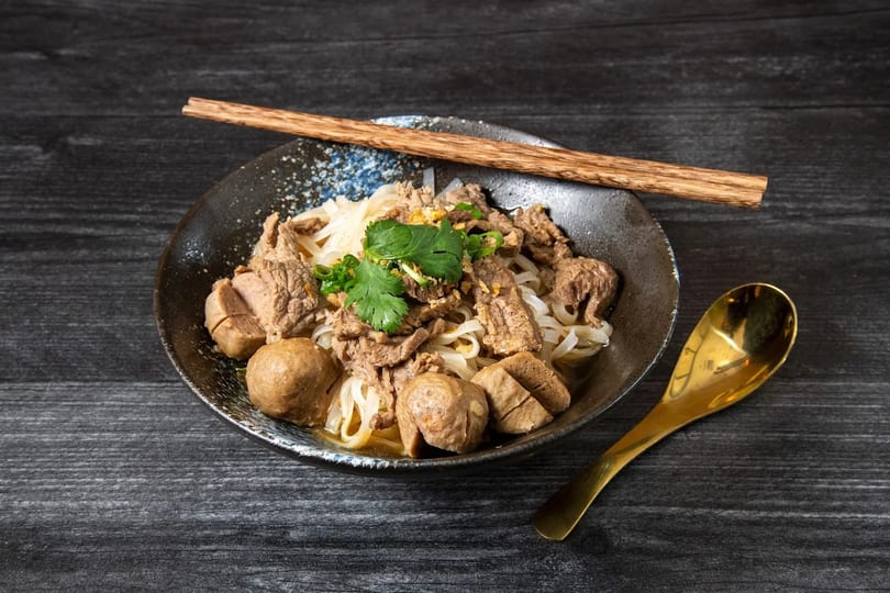 A bowl of Thai boat noodles with beef, meatballs, and cilantro, with chopsticks and a spoon.