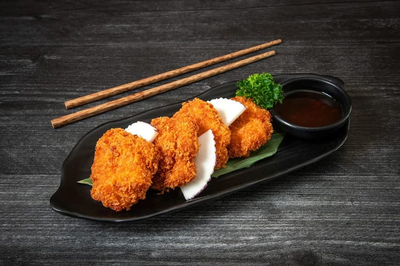 Crispy fried shrimp cakes with radish slices, parsley, and dipping sauce on a black plate with chopsticks.