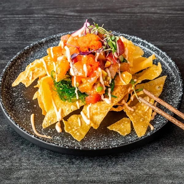 Poke nachos with salmon, tuna, seaweed, roe, and sauce on a dark plate with chopsticks.