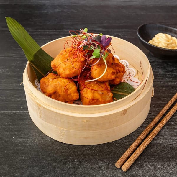 Fried chicken pieces in a bamboo steamer with chopsticks and a dipping sauce.