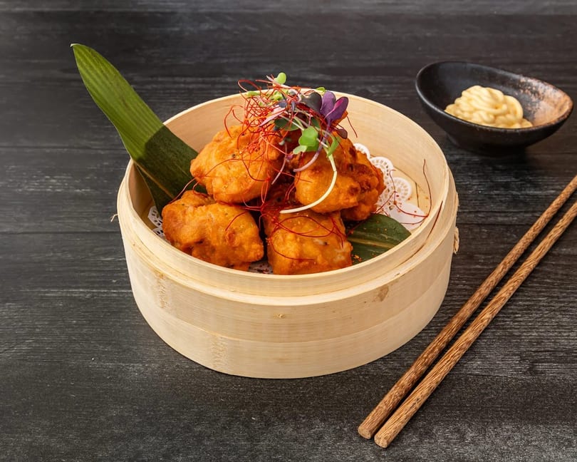 Fried chicken pieces in a bamboo steamer with chopsticks and a dipping sauce.