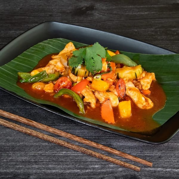 Sweet and sour chicken with vegetables on a banana leaf, served on a black plate with chopsticks.