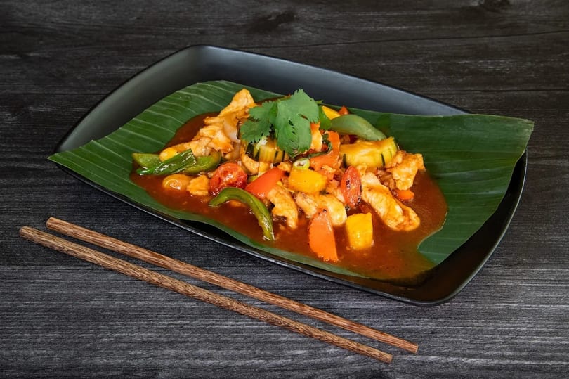 Sweet and sour chicken with vegetables on a banana leaf, served on a black plate with chopsticks.