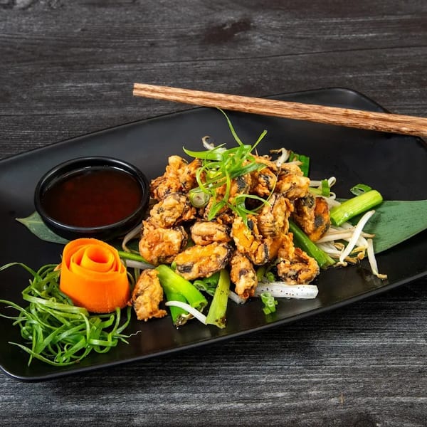 Crispy fried mussels with green onions, bean sprouts, carrot rose, and dipping sauce on a black plate.