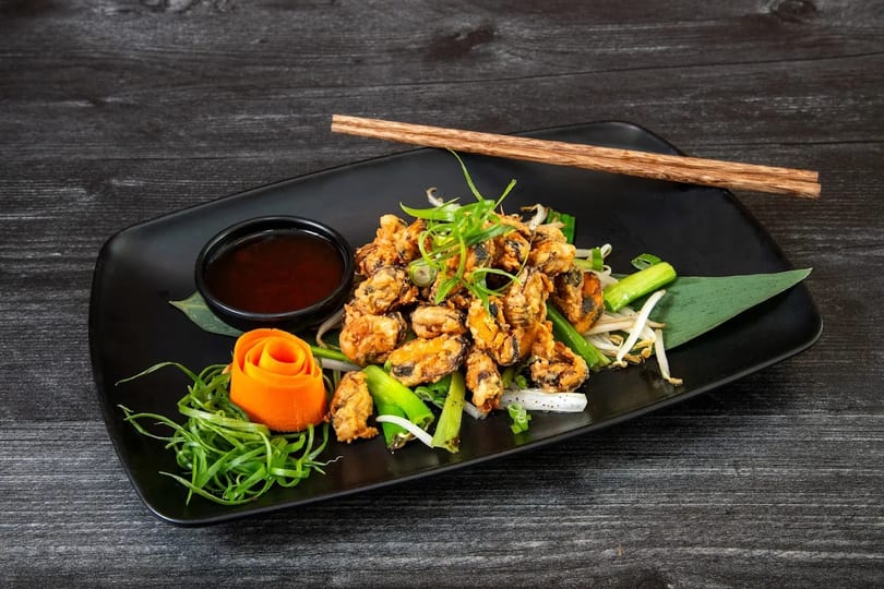 Crispy fried mussels with green onions, bean sprouts, carrot rose, and dipping sauce on a black plate.
