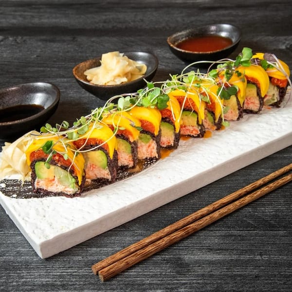 Sushi roll with mango, avocado, and crab, garnished with microgreens, on a white plate with chopsticks.