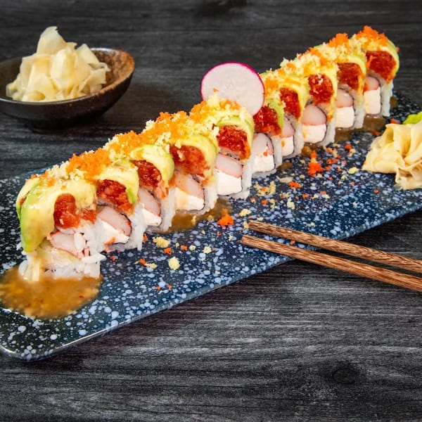 Sushi roll with avocado, red topping, and orange roe, served with ginger and chopsticks.