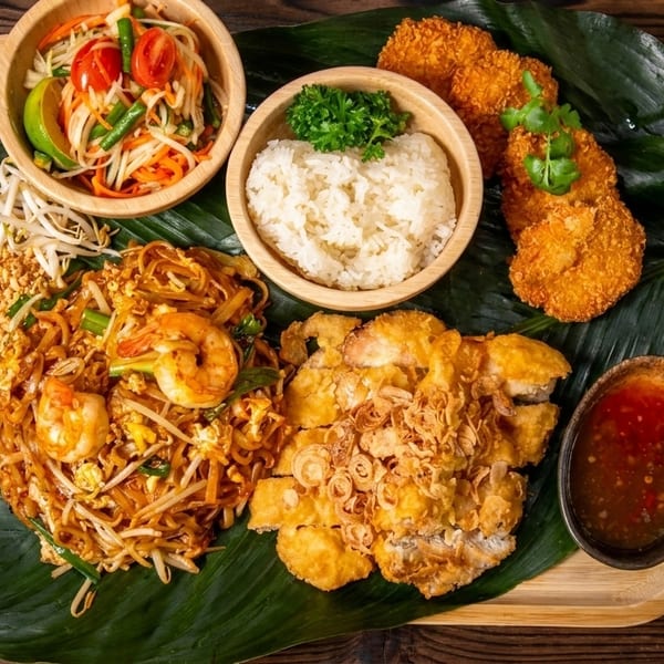 A wooden tray with various Thai dishes including Pad Thai, papaya salad, rice, and fried chicken.