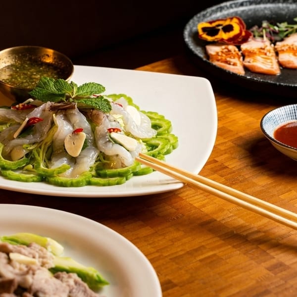 A table spread with various Asian dishes, including shrimp, salmon, and pork, with chopsticks and dipping sauces.